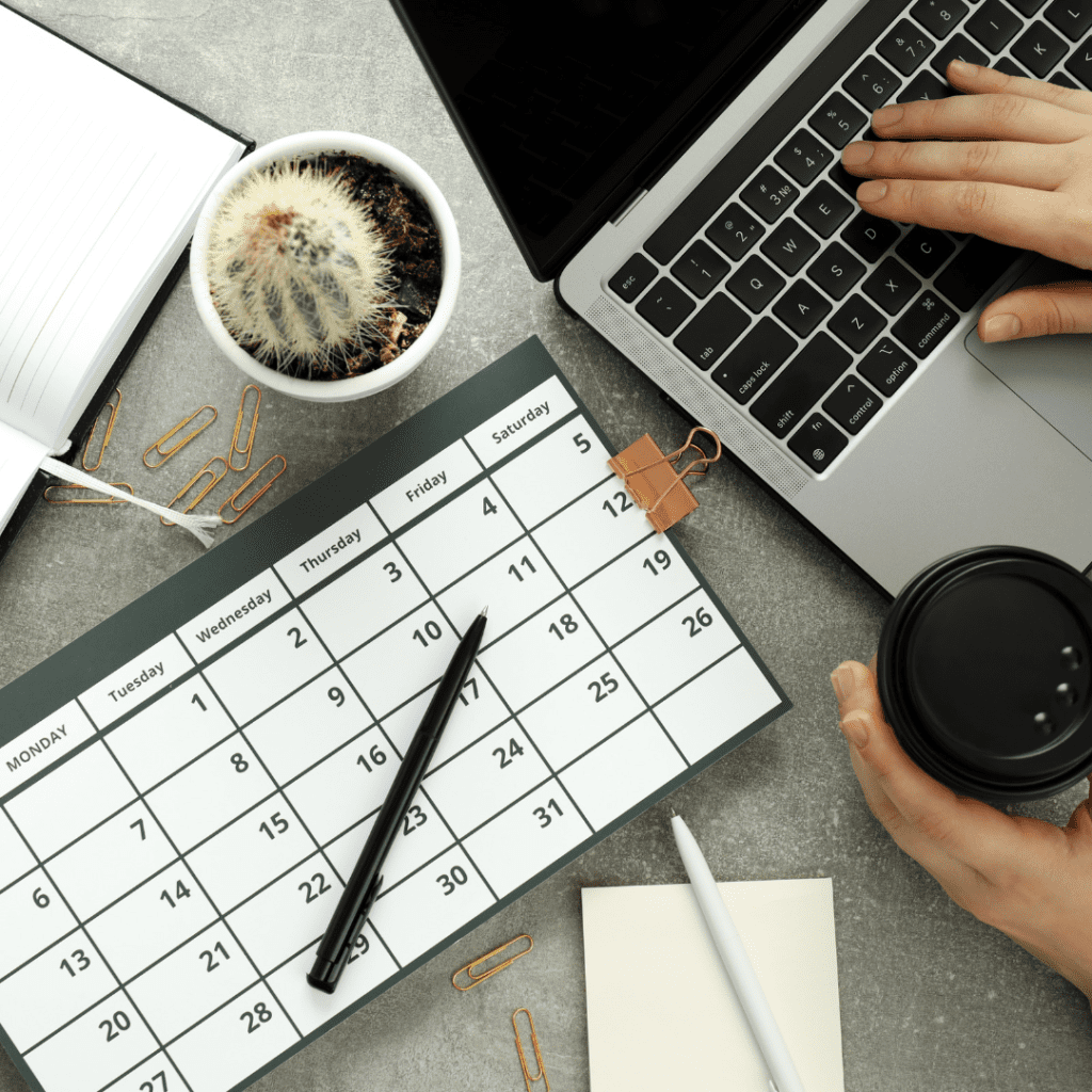 A person's hand typing on a laptop keyboard while the other hand holds a black coffee cup. The desk also has a calendar open to tips for hiring and working with contractors, an open notebook, a pen, paper clips, a small cactus plant, and sticky notes.